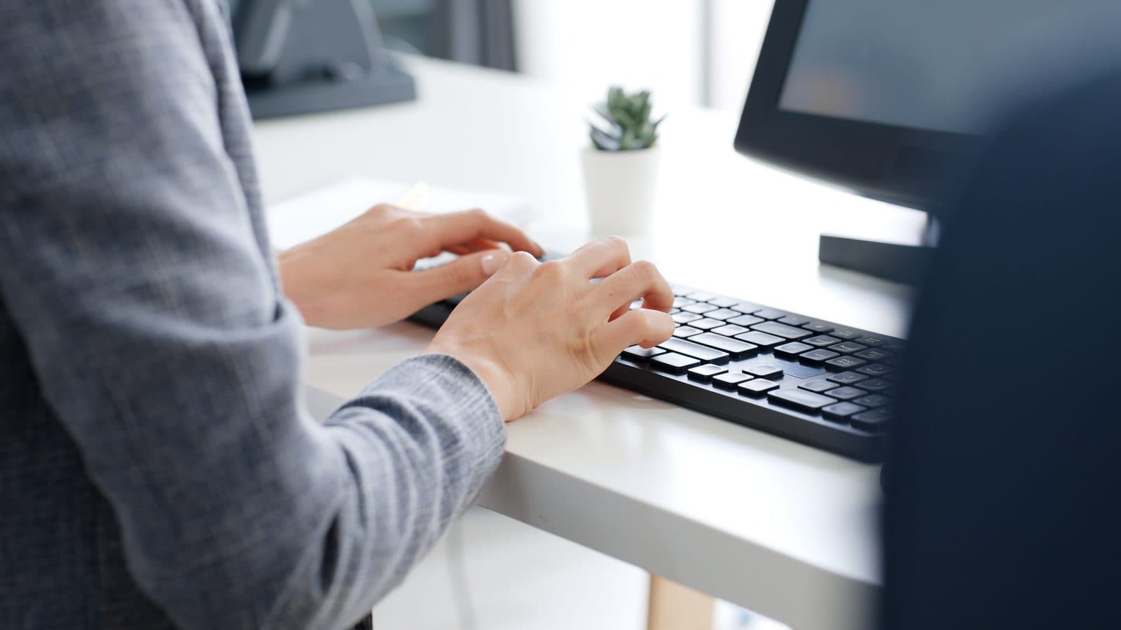 Hands typing on a keyboard at a white desk with a monitor and a small potted plant