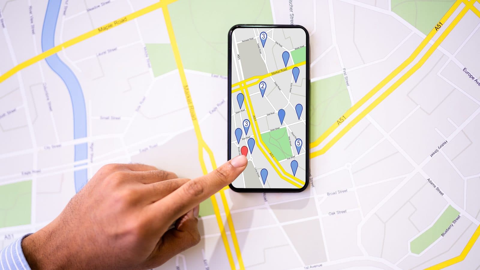 A finger tapping a red pin on a phone map, with the phone resting on a paper street map